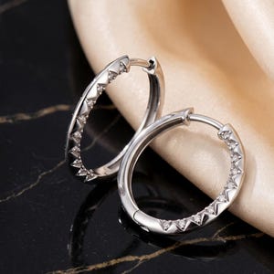 May include: A pair of silver hoop earrings with a row of small, sparkling gemstones set along the outer edge. The earrings are displayed on a black surface with a gold-colored fabric in the background. The earrings have a polished, reflective finish.