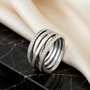 May include: A silver ring featuring multiple intertwined bands, some set with small, clear gemstones. The ring has a contemporary design and is presented on a black and gold marble surface with a white satin backdrop.