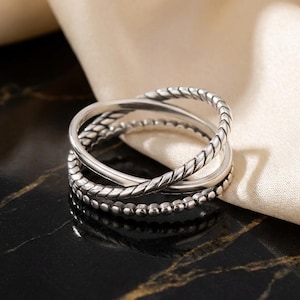 May include: A silver ring set with multiple bands. The bands have varied textures, including a twisted rope design, a smooth band, and a band with small beaded details. The ring is shown on a black surface with gold veining.
