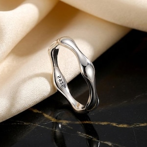 May include: A silver ring with a fluid, organic design. The ring features a wavy shape with a smooth, polished surface. The inside of the ring is engraved with the number "2352". The ring is presented on a cream-coloured fabric and a black surface.