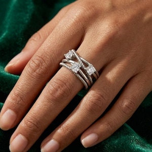 May include: A silver ring featuring three clear gemstones. The ring has a twisted rope design and is worn on a finger. The ring is set against a dark green background.