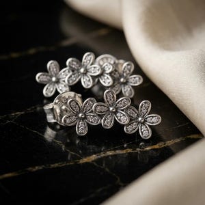 May include: A pair of silver-toned ear climbers featuring a cluster of three connected flower shapes. Each flower petal is encrusted with small, clear, sparkling stones. The earrings have a post-back closure and a polished metallic finish.