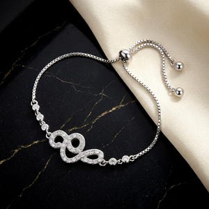 May include: A silver-toned adjustable bolo bracelet featuring a central infinity-inspired motif encrusted with small clear stones. The chain is a delicate box link style, accented by three small round stones on each side and two polished metal beads.