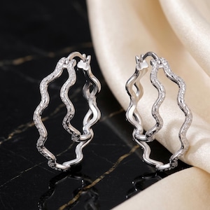 Può includere: Un paio di orecchini a cerchio argentati con un design ondulato. Gli orecchini sono adornati con piccole pietre scintillanti, che aggiungono un tocco di eleganza. Sono esposti su una superficie nera con un tessuto color crema sullo sfondo.