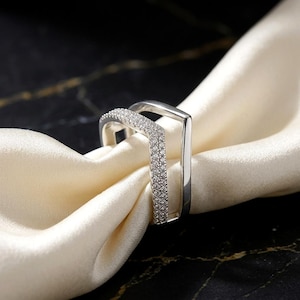 May include: A silver-toned V-shaped ring featuring a double-band design. One band is encrusted with numerous small, clear, sparkling stones, while the other band is a smooth, polished, solid silver-toned metal. The ring has a sharp, angular silhouette.