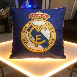 May include: A square throw pillow featuring a deep blue background with the Real Madrid football club logo printed in the center. The emblem displays a gold crown above a circular crest containing the letters MCF in gold, white, and blue.