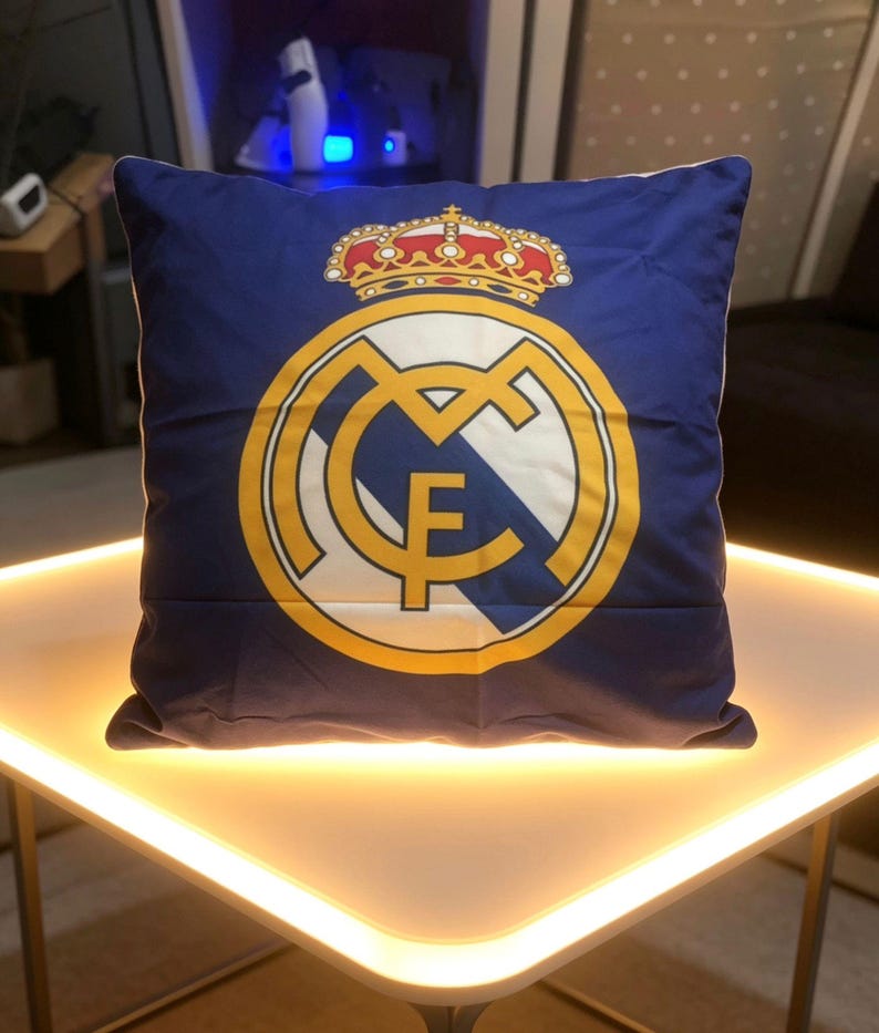 May include: A square throw pillow featuring a deep blue background with the Real Madrid football club logo printed in the center. The emblem displays a gold crown above a circular crest containing the letters MCF in gold, white, and blue.