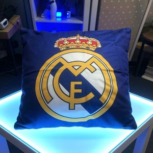 May include: A square blue throw pillow featuring the Real Madrid soccer club logo printed in the center. The design includes a large yellow and white circular emblem with the letters MCF and a detailed red, yellow, and white crown on top.
