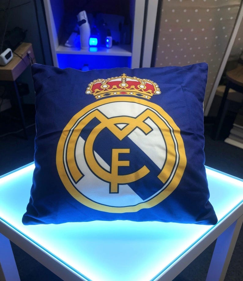 May include: A square blue throw pillow featuring the Real Madrid soccer club logo printed in the center. The design includes a large yellow and white circular emblem with the letters MCF and a detailed red, yellow, and white crown on top.