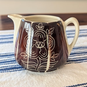 May include: A vintage ceramic creamer with a brown and cream color scheme. The creamer features a decorative floral design in white against a brown background. It has a spout and a handle, and is placed on a blue and white striped cloth.