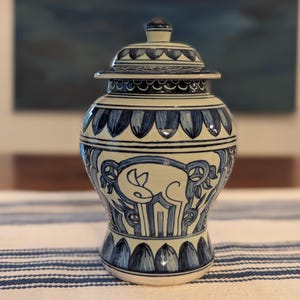 May include: A ceramic ginger jar with a lid, featuring a cream-colored base and hand-painted dark blue patterns. The design includes repeating petal motifs around the neck and base, with stylized animal figures depicted on the main body.
