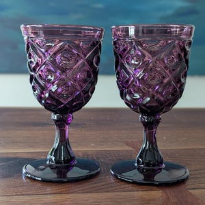 May include: Two matching purple glass goblets featuring a raised diamond and circular pattern on the bowl. Each goblet has a short, thick stem and a wide, circular base, crafted from translucent deep purple glass with a smooth, polished finish.