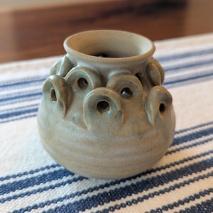 May include: A small, round, light green ceramic vase with a speckled finish. The vase features a wide opening at the top and a series of decorative, arched loops or small handles arranged in a ring around the upper body, creating multiple openings.