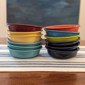 May include: Two stacks of ceramic bowls featuring concentric ring patterns along the exterior rims. The left stack includes deep red, yellow, and turquoise bowls. The right stack features orange, blue, lime green, and dark navy blue bowls.