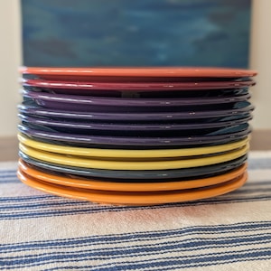 May include: A stack of round ceramic plates in a variety of colors including orange, dark red, purple, dark blue, yellow, and grey. The plates have a smooth, glossy finish and are stacked vertically, showing the edges of each individual plate.
