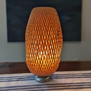 May include: A woven rattan table lamp with a warm, amber glow. The lamp has a cylindrical shape with a diamond pattern and sits on a silver-colored base. The lamp is on a table with a blue and white striped runner.