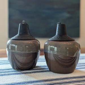 May include: Two ceramic oil lamps with dark brown and gray glaze. Each lamp has a black lid with a chain and the text "CMC". The lamps are on a striped tablecloth.