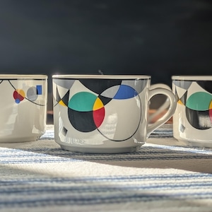 May include: Three white ceramic mugs featuring a geometric pattern of black lines, circles, and shapes in red, yellow, blue, and teal. Each mug has a smooth cylindrical body, a curved handle on the side, and a base that is slightly recessed.