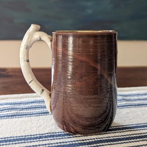 May include: A cylindrical ceramic mug with a glossy dark brown glaze featuring horizontal ridges along the body. The handle is shaped like a white birch tree branch with small dark gray markings, contrasting with the smooth, dark brown finish of the mug.