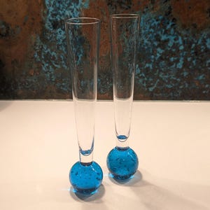 May include: Two clear glass vases with long, slender stems and round, blue bases. The bases are decorated with small, bubble-like inclusions. The vases are displayed on a white surface against a textured, blue and brown background.