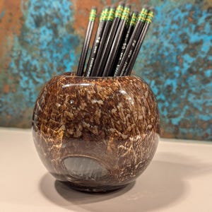 May include: A brown and white speckled glass pencil holder with a round shape. The pencil holder contains several black pencils with green and yellow accents. The pencils are arranged upright in the holder. The background is a textured blue and brown.