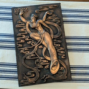 May include: A rectangular wall plaque featuring a raised copper-toned relief of a celestial figure in motion. The figure is surrounded by stylized cloud motifs against a dark, textured background. The design showcases intricate flowing garments.