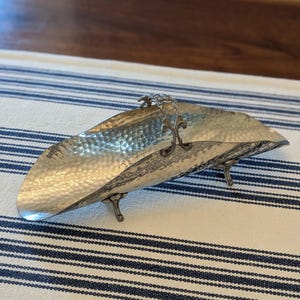 May include: A silver-toned, elongated, boat-shaped serving dish with a hammered metal texture. The dish features a central decorative arched handle with intricate scrollwork and is supported by three small, ornate metal feet.