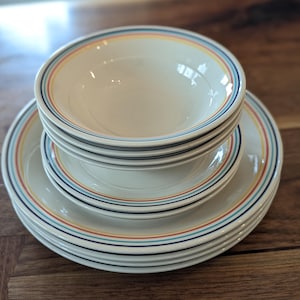 May include: A stack of off-white ceramic bowls and plates. Each piece has a colorful striped border in blue, red, yellow, and black. The bowls are stacked on top of the plates, creating a cohesive dinnerware set.