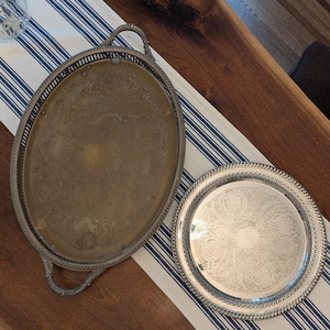 May include: An oval brass-toned serving tray with ornate handles and a circular silver-toned serving tray, both featuring intricate engraved patterns and decorative pierced lattice borders along the edges of the metal surfaces.