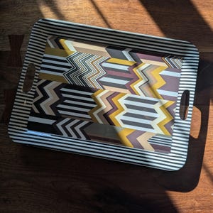 May include: A rectangular serving tray featuring a black and white striped border and two integrated side handles. The center surface displays a repeating geometric pattern of zig-zagging lines in shades of mustard yellow, dark brown, and light gray.