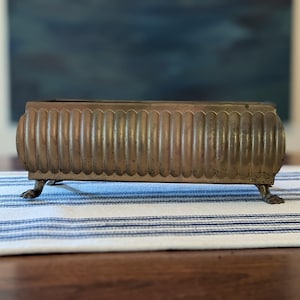May include: A rectangular brass planter featuring a vertical fluted pattern along the exterior walls. The vessel is supported by four small, detailed claw-style feet at each corner, showcasing a weathered metallic finish with visible patina.