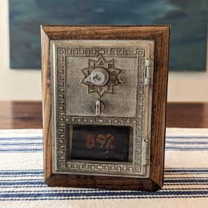 May include: A vintage post office box door set in a dark wood frame. The silver-toned metal door features a star-shaped dial with letters A through J, a central latch, and a glass window displaying the red painted number 692 with a Greek key border.