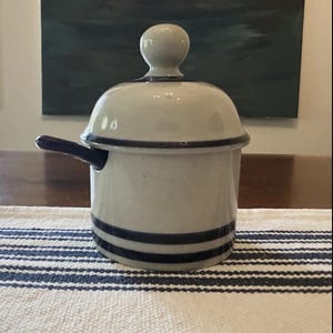 May include: A cylindrical ceramic jar with a matching domed lid featuring a knob handle. The jar is off-white with two dark blue horizontal stripes around the base and a single dark blue stripe around the lid. A dark blue handle extends from the side.