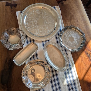 May include: An assortment of antique silver serving trays and dishes. The collection includes round, oval, and rectangular shapes with ornate embossed designs and decorative edges. The items are arranged on a striped cloth and a wooden surface.