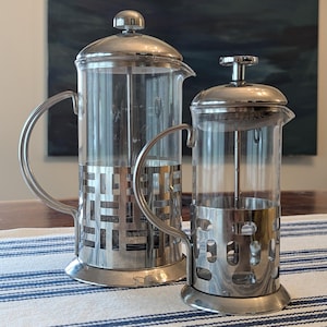 May include: Two stainless steel and glass French presses of different sizes. The presses have a cylindrical glass body, a metal filter, and a lid. The larger press has a decorative metal band around the base. The handles are curved.