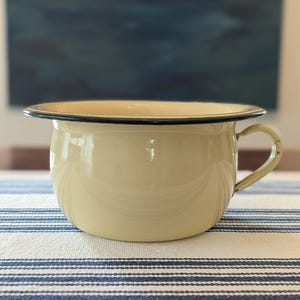 May include: A cream-colored enamel chamber pot featuring a wide, rounded basin, a smooth glossy finish, and a single curved handle on the side. The rim of the basin is accented with a thin, dark black border that contrasts with the light body.