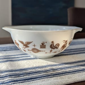 May include: White mixing bowl with brown decorative images of a rooster, eagle, and other kitchen-themed illustrations. The bowl has a wide rim and is sitting on a blue and white striped tablecloth.