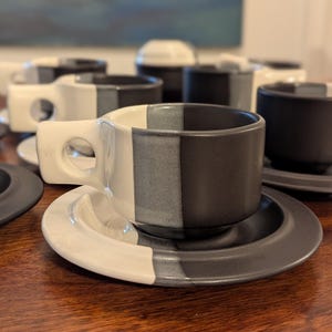 May include: A ceramic cup and saucer set featuring a two-tone design with a glossy white finish on one side and a matte black finish on the other. The cup includes a distinctive handle with a circular cutout, and the saucer matches the color scheme.