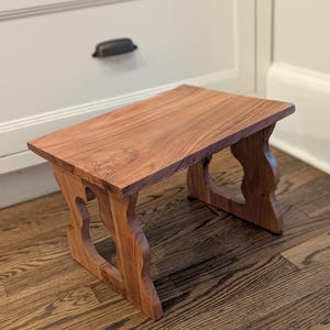 May include: A small, handcrafted wooden step stool. The stool is made of reddish-brown wood with a rectangular top and decorative, curved legs. The legs feature heart-shaped cutouts. The stool is designed for use as a step or a decorative accent.
