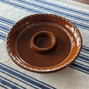 May include: A brown ceramic serving dish with a central raised section and a decorative white scalloped edge. The dish is set on a blue and white striped cloth, showcasing its rustic design. This piece is ideal for serving dips or sauces.