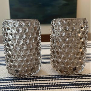 May include: Two cylindrical glass tumblers featuring a distinct hobnail texture with raised, rounded bumps across the entire exterior surface. The glass has a translucent, smoky grey tint, creating a uniform pattern of light and shadow.