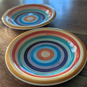 May include: Two ceramic bowls with concentric rainbow-colored circles. The bowls have a shallow design with a cream-colored rim. The color pattern includes orange, red, yellow, green, blue, and purple.