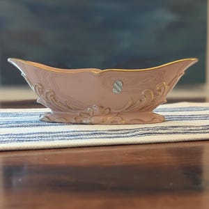 May include: A pink ceramic decorative bowl with gold trim and ornate detailing. The bowl has a boat-like shape with flared sides and a scalloped rim. A small label is affixed to the side. The bowl is resting on a striped fabric.