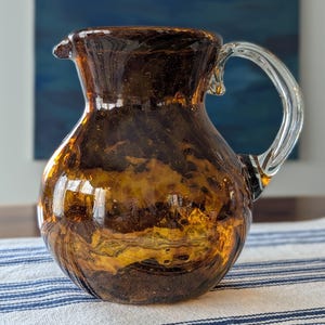 May include: A decorative amber glass pitcher with a clear glass handle. The pitcher has a spout and a rounded body. The glass has a marbled appearance with shades of brown and gold. The pitcher is sitting on a blue and white striped tablecloth.