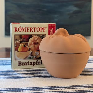 May include: A terracotta apple-shaped baking dish with a lid, featuring the embossed text RÖMERTOPF on the top. Beside it stands a cardboard box labeled RÖMERTOPF and Bratapfel, displaying an image of a baked apple inside the same dish.