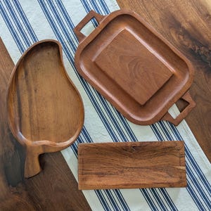 May include: A collection of wooden serving trays in various shapes. The trays include a leaf-shaped tray, a rectangular tray, and a tray with handles. The wood has a warm, brown tone, and the trays are arranged on a striped blue and white cloth.