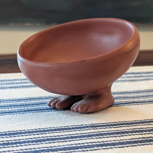 May include: A terracotta-colored bowl with a rounded shape, supported by two feet. The bowl has a smooth surface and is set on a blue and white striped tablecloth. The feet are detailed with toes.