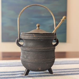 May include: A dark gray cast iron pot with three short legs, two side handles, and a matching lid. A brass-colored metal handle arches over the top, and a long, thin brass-colored metal spoon rests inside the pot, protruding from the lid opening.