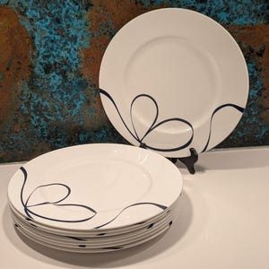 May include: A stack of white ceramic plates with a navy blue abstract design. One plate is displayed on a black stand. The plates have a simple, elegant design, suitable for everyday use or special occasions.