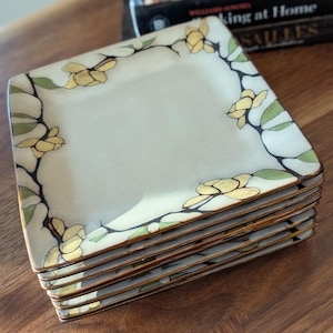 May include: A stack of square plates with a cream-colored surface. Each plate is decorated with a floral design featuring yellow flowers, green leaves, and black stems around the edges. The plates are stacked on a wooden surface.